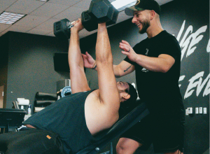 Personal trainer assisting a client with dumbbell strength training in a gym in Thane.